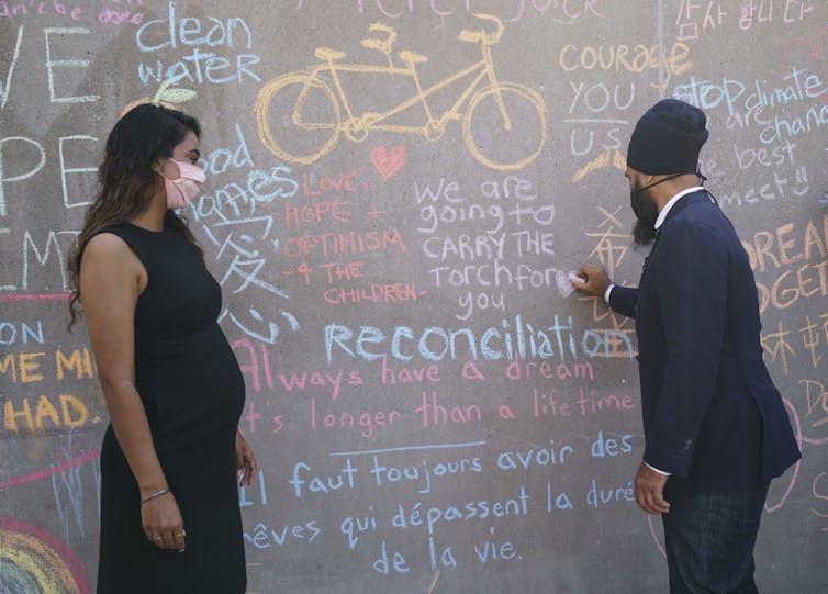 Jagmeet Singh writing with chalk on a wall at a news conference as his wife Gurkiran Kaur Sidhu looks on