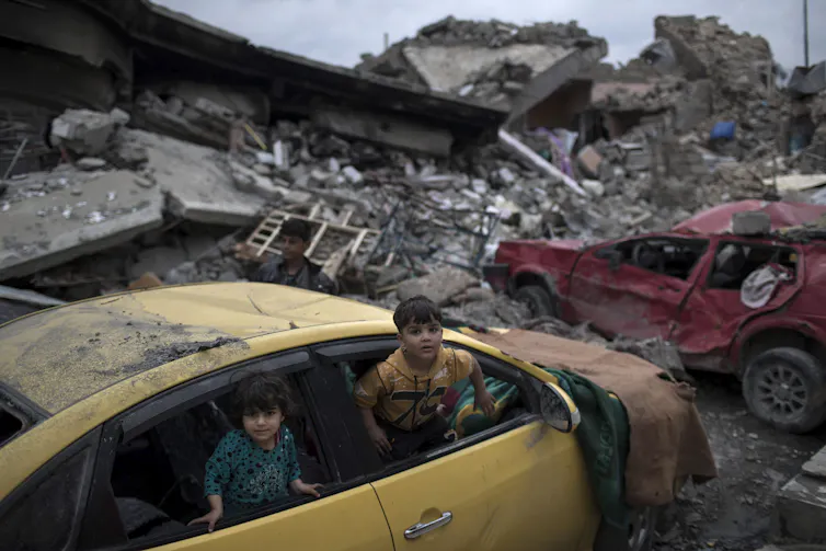 Children play in a damaged car amid piles of rubble.