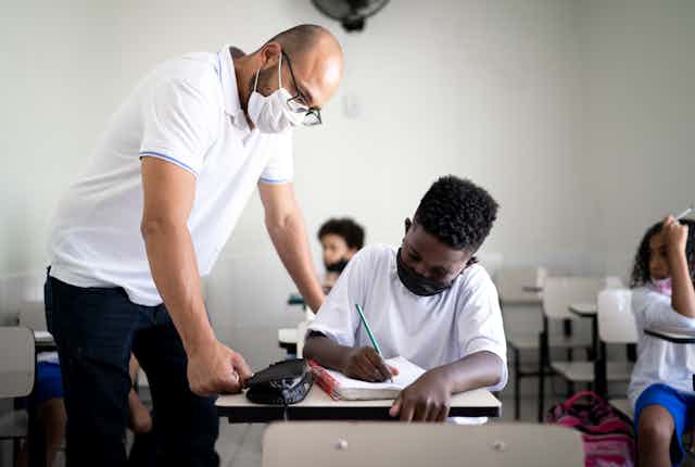 A mask-wearing teacher helps a Black student, who is also wearing a mask, on his classwork.