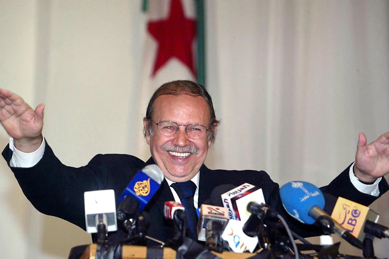 A man on grey suit, speaking with his hands raised