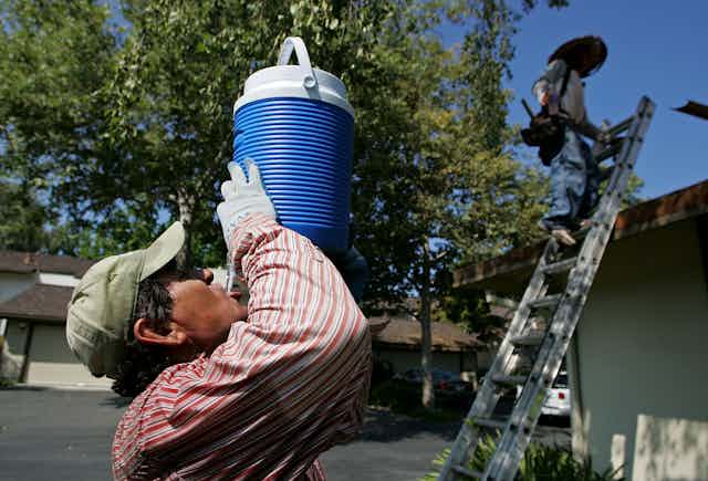 A sweating roofer drinks cold water from a container with a home in the background