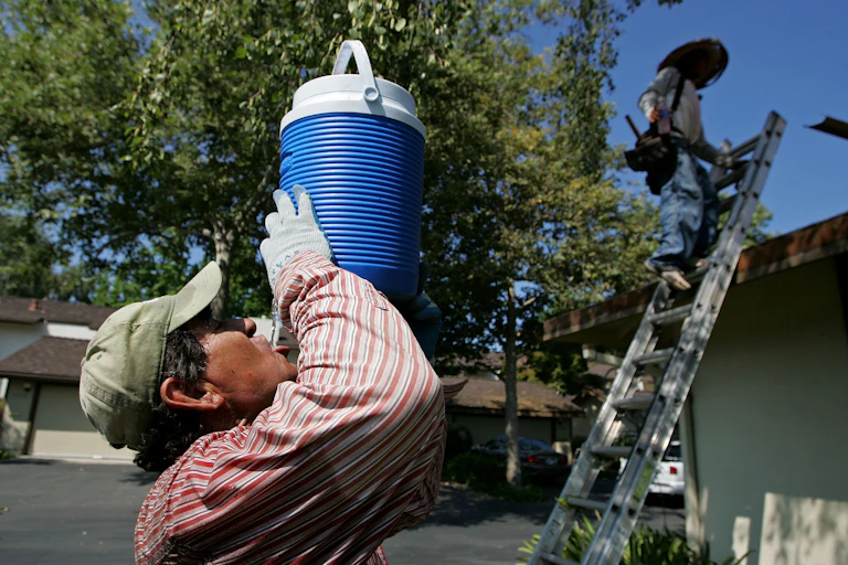A sweating roofer drinks cold water from a container with a home in the background
