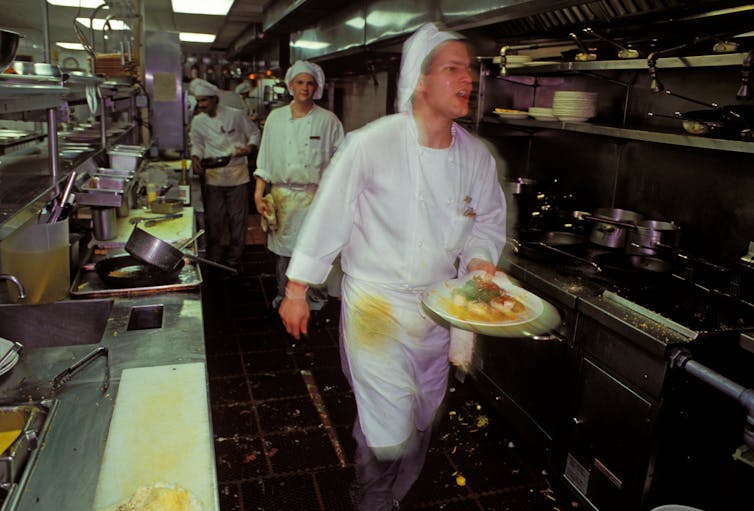 A sweating cook carries as plate past rows of ovens. The photo is shot with the cook slightly blurred, capturing the frenetic pace of the kitchen