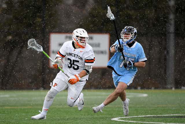 Two young white athletes play lacrosse on a field.