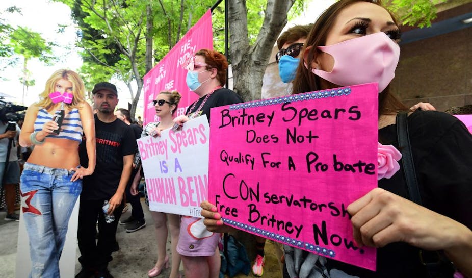 A woman with brown hair wearing black and pink holds a pink sign that reads, "Britney Spears Does Not Qualify For A Probate CONservatorship. Free Britney Now!" In the background, four fans are also gathered in protest with a large cutout of Britney Spears