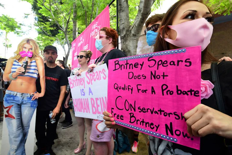 A woman with brown hair wearing black and pink holds a pink sign that reads, "Britney Spears Does Not Qualify For A Probate CONservatorship. Free Britney Now!" In the background, four fans are also gathered in protest with a large cutout of Britney Spears