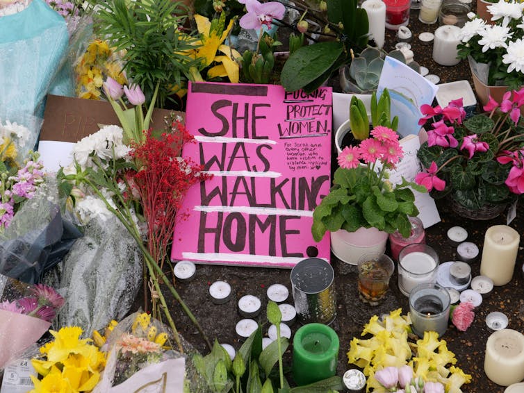 A pink sign with the handwritten message 'She was walking home'' on the ground surrounded by flowers and candles