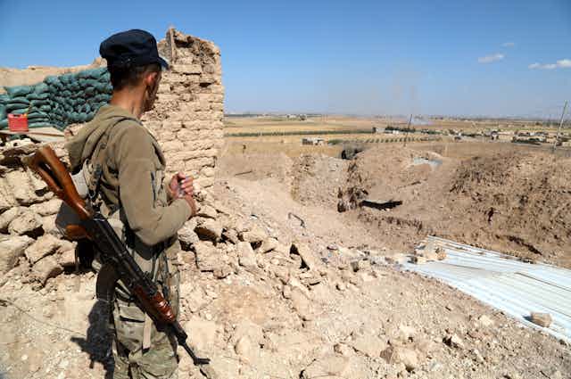 A soldier with an automatic rifle stands by a defensive encampment looking at Turkish troops in the distance.