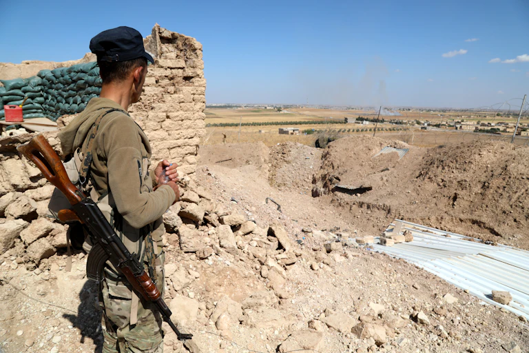 A soldier with an automatic rifle stands by a defensive encampment looking at Turkish troops in the distance.