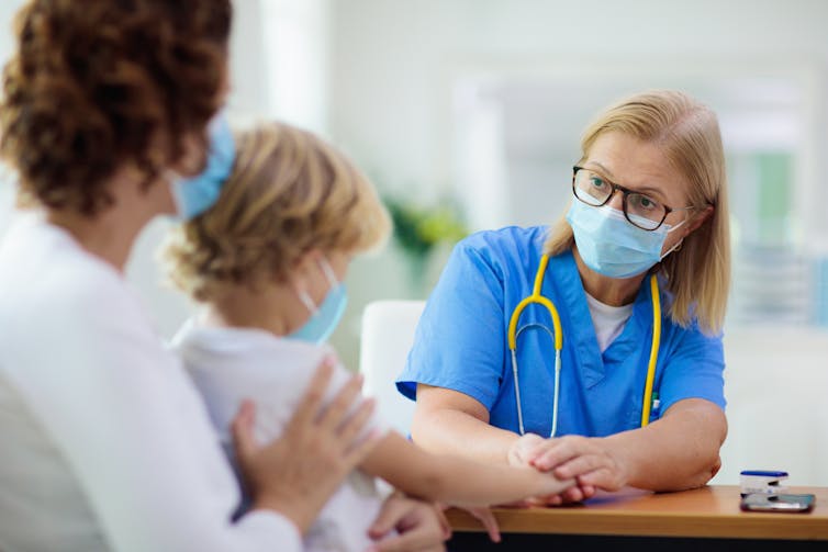A doctor wearing blue scrubs and a mask examines a child sitting on their mother's lap
