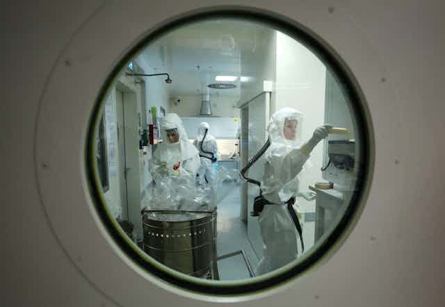 Looking through a porthole type window into a lab. Three workers are in full body PPE.