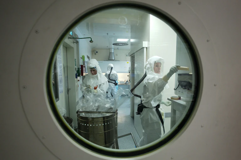 Looking through a porthole type window into a lab. Three workers are in full body PPE.
