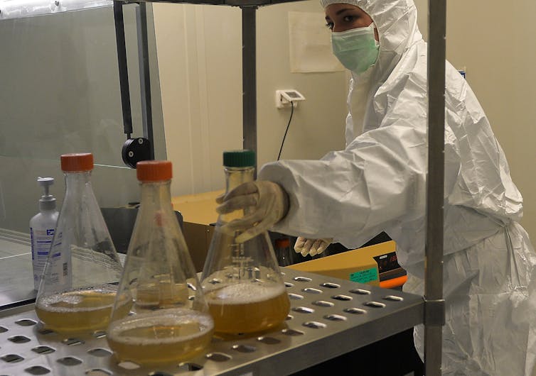 A woman in full body PPE reaches for a beaker containing a murky liquid.