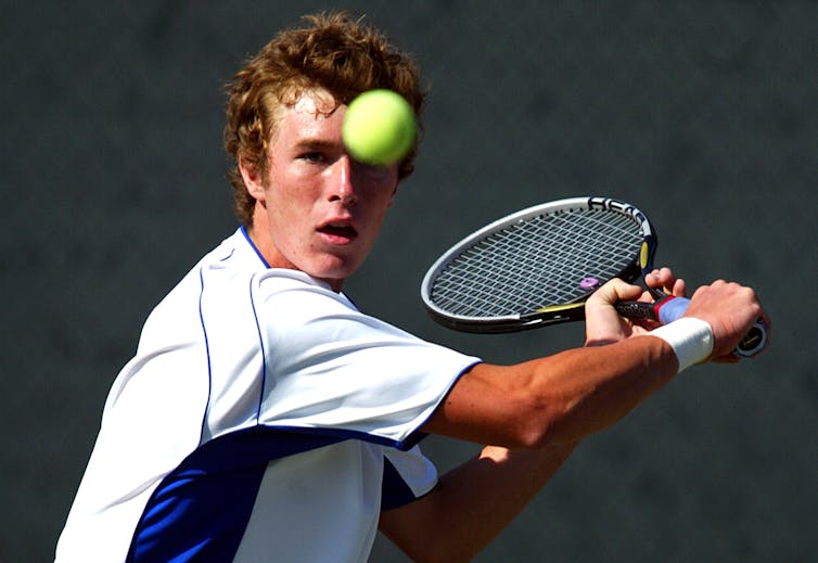 A young white man attempts to hit a tennis ball with a racket.