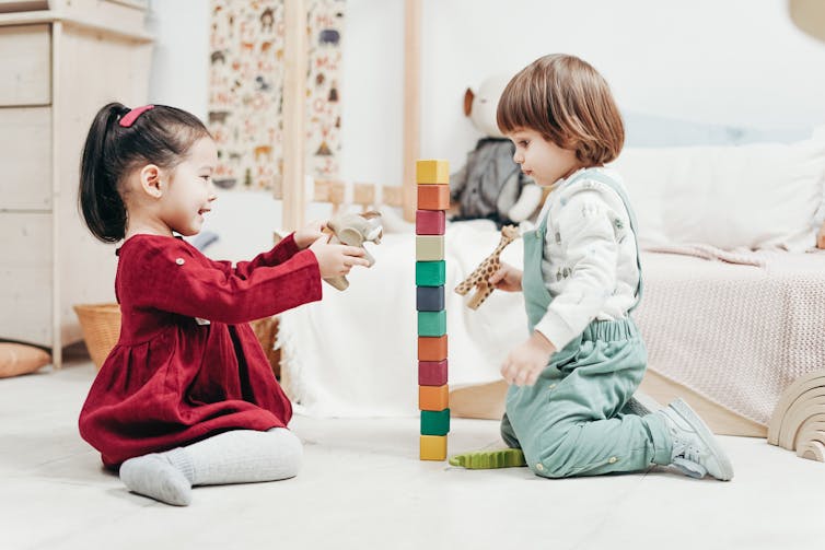 Two preschoolers play with blocks, one holding an elephant and one holding a giraffe, while sitting on the floor.