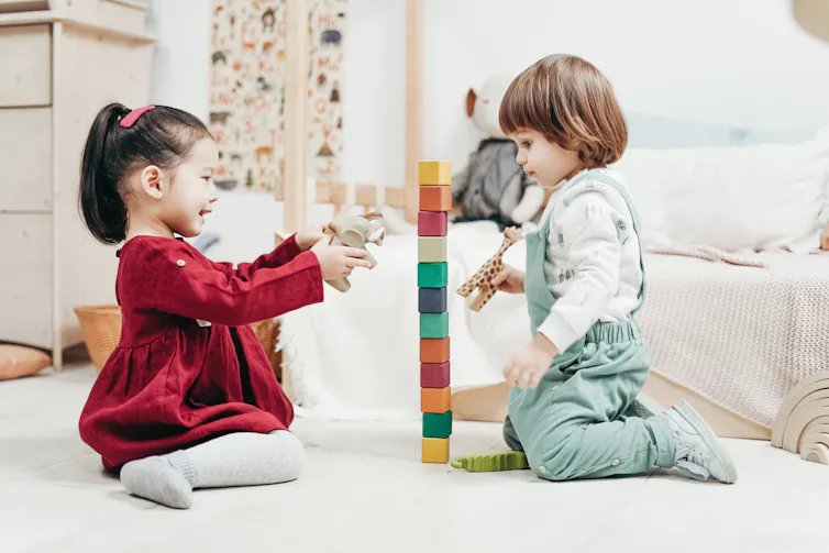 Two preschoolers play with blocks, one holding an elephant and one holding a giraffe, while sitting on the floor.