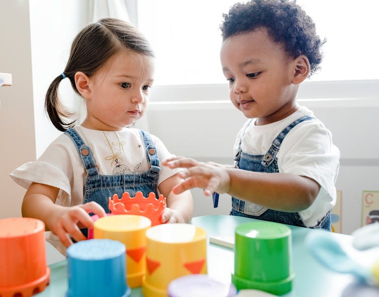 Two preschooler children in overalls play with stackable cups.