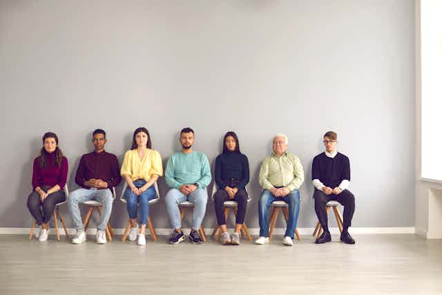 A row of men and women sit on stools outside a room.