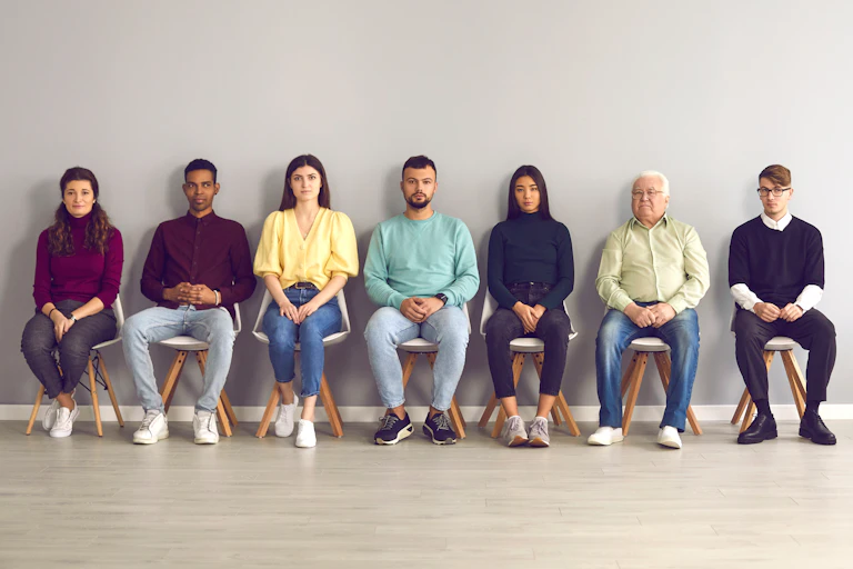 A row of men and women sit on stools outside a room.