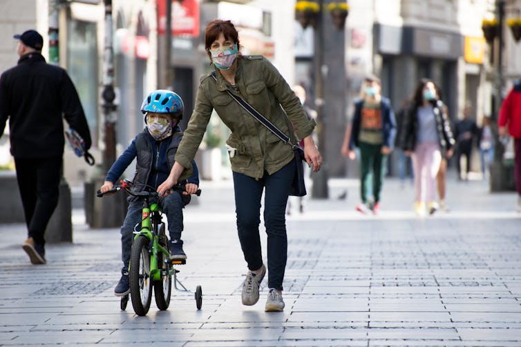 A mother walking on a paved street, helps her child who is riding a bike.