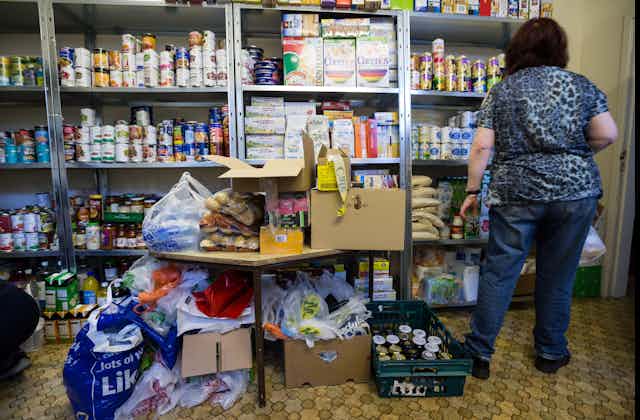 A woman volunteering in a food bank. She has her back to the camera and is facing shelves stacked with tins and boxes of food.