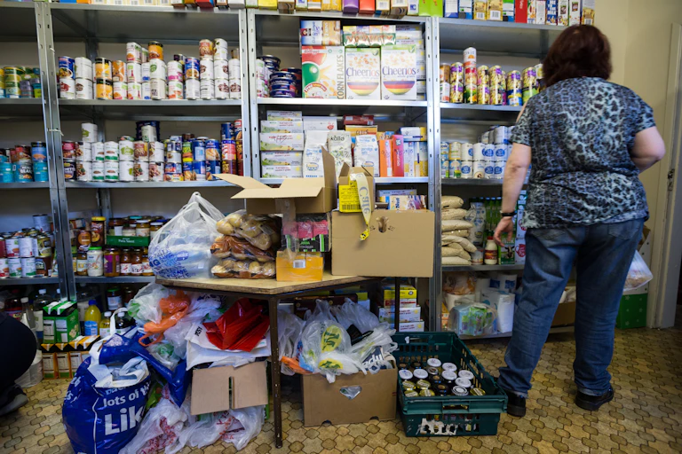 A woman volunteering in a food bank. She has her back to the camera and is facing shelves stacked with tins and boxes of food.