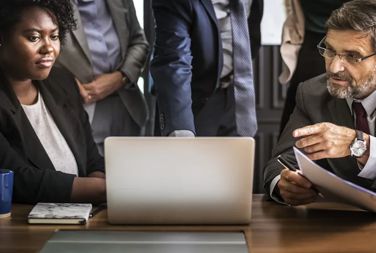 People looking stern-faced gather around a laptop in an office setting.