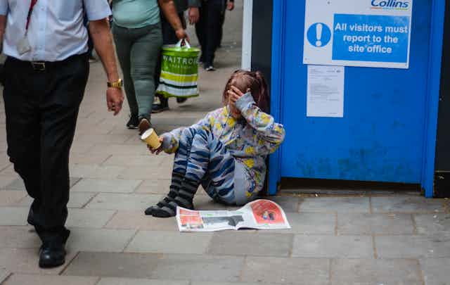 A young girl sits on a sidewalk in London with a cup, asking for money. She has no shoes on and has her hand over her eyes as people walk past.