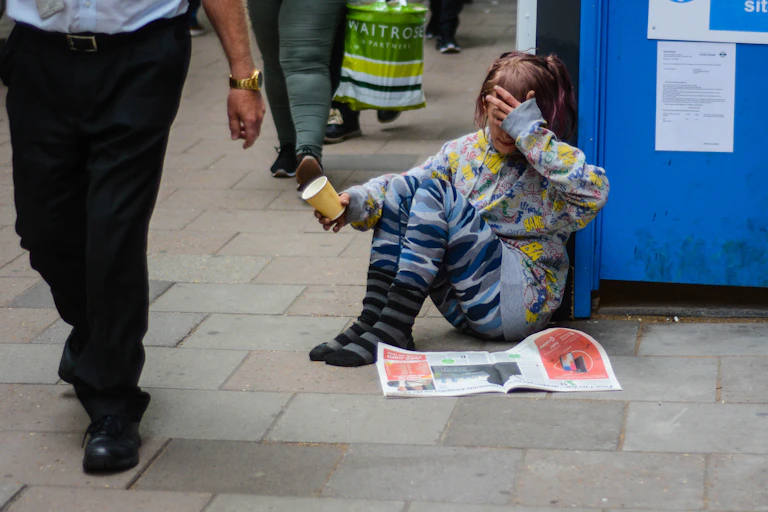 A young girl sits on a sidewalk in London with a cup, asking for money. She has no shoes on and has her hand over her eyes as people walk past.