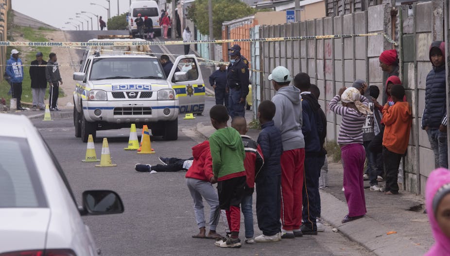 Children watch as police investigate at a scene where a young man lies dead on the road