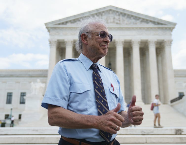 Older man in a short-sleeved dress shirt, tie and sun glasses with two thumbs up