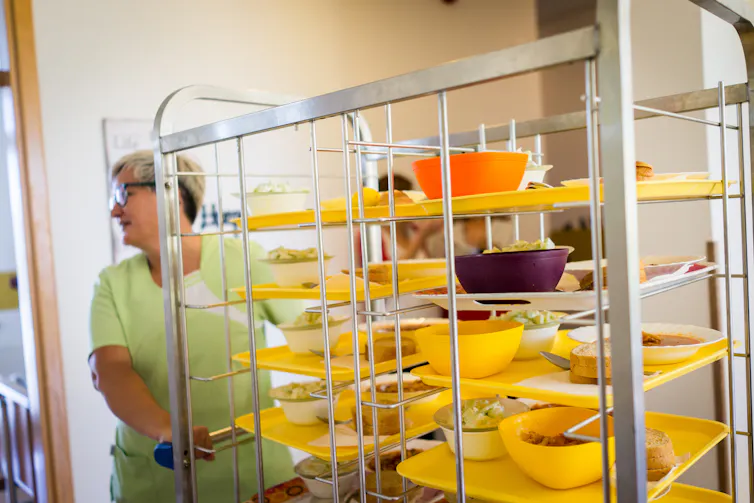 Person in scrubs wheeling bowls of food in stacked on a cart in a nursing home.