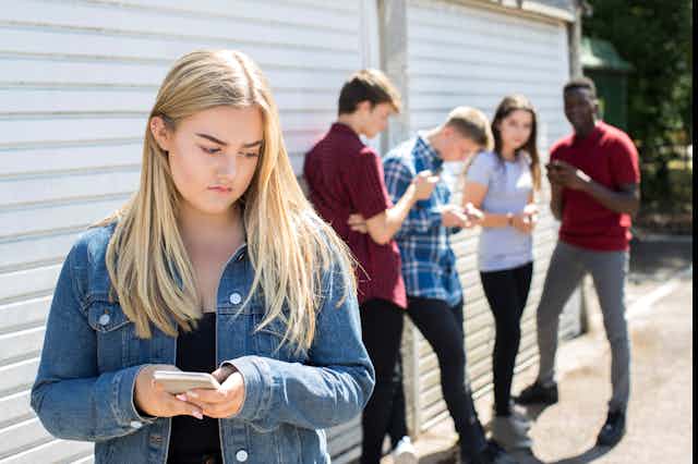 A teenage girl holding a smartphone, looks unhappy. Other teenagers stand in the background.