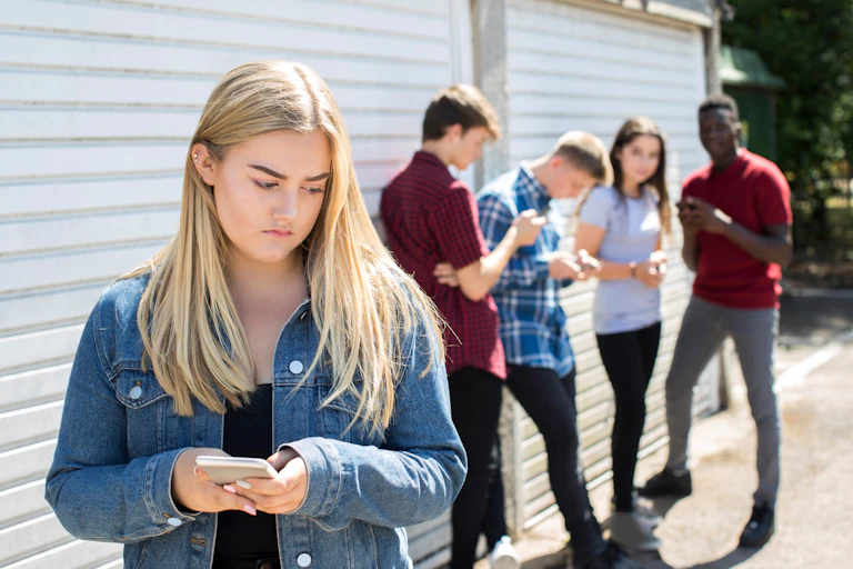 A teenage girl holding a smartphone, looks unhappy. Other teenagers stand in the background.