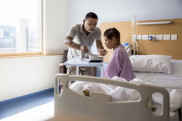 Person feeding an older patient sitting on a hospital bed.