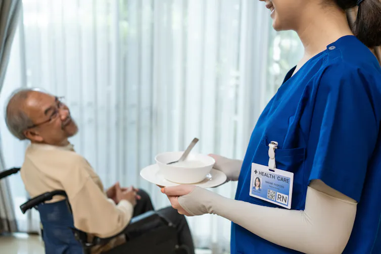 Nurse offering a bowl of food to an older adult in a wheelchair.