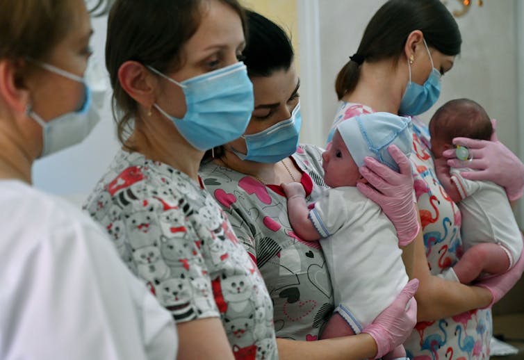 Four women standing together wearing masks, with two of them holding new-born babies.