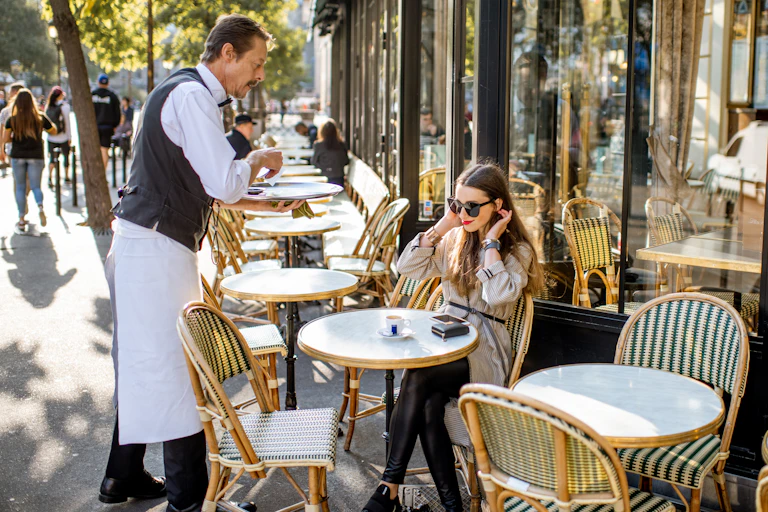 Waiter serving coffee to a young woman client sitting at the traditional french cafe outdoors in Paris.