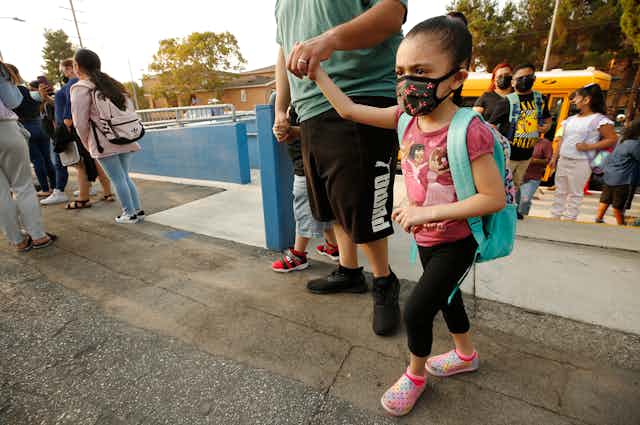 Man holds little girl's hand as she walks into school wearing a backpack and face mask