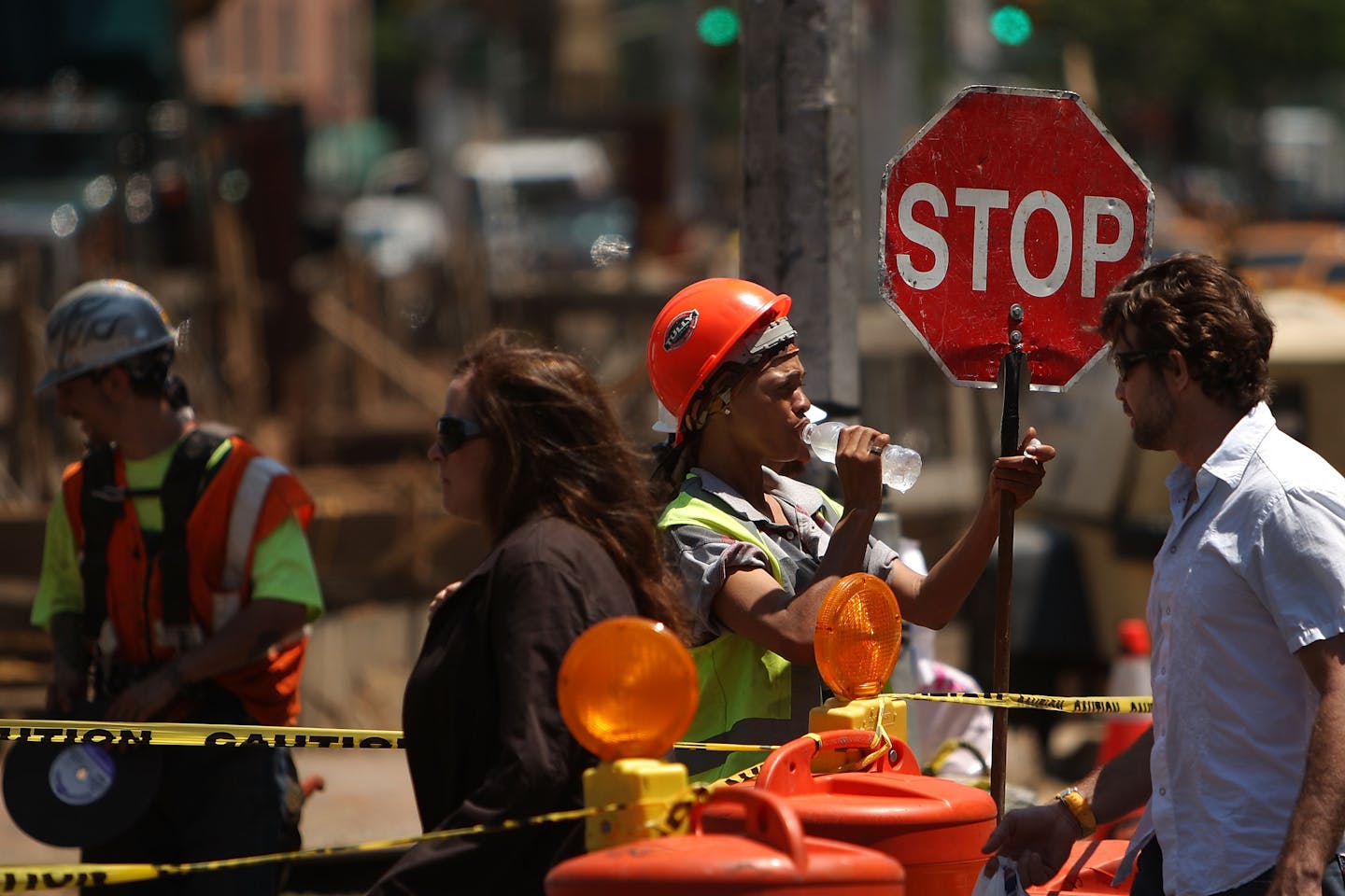 A construction worker, in a hard hat and drinking from a water bottle, holds a stop sign as people cross the street on a hot day in New York City.