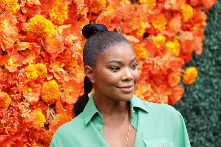 A woman in a green shirt stands in front of colorful red and orange flowers.