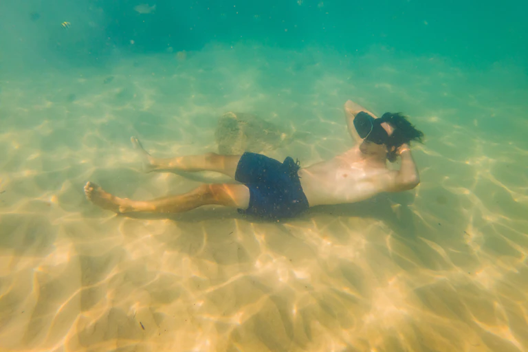 A man wearing blue swim trunks and a VR set floating on a seabed