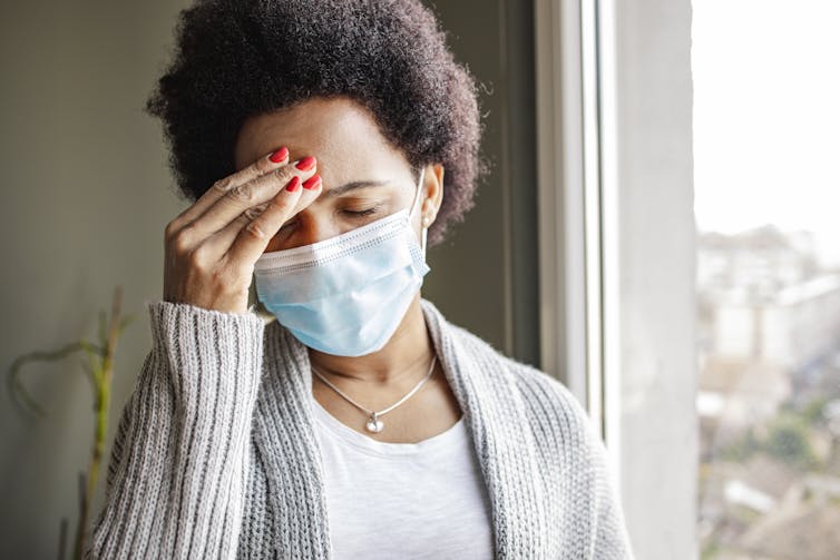 An African American woman looking stressed and wearing a face mask by a window.