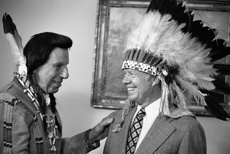 A man dressed in Native American clothes, Iron Eyes Cody, giving President Jimmy Carter a Native American headdress. Neither man was Native American.