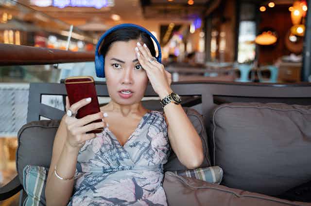 a seated woman wearing headphones and holding a smartphone in her right hand presses her left hand to her forehead with an exasperated expression on her face