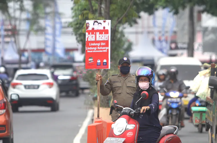 Petugas berdiri menunjukkan poster imbauan pencegahan COVID-19 di tepi jalan.