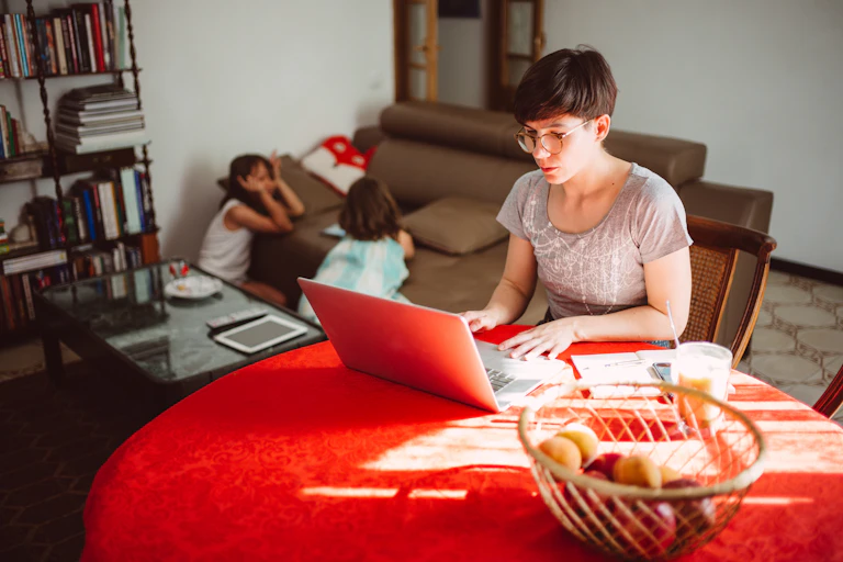 A woman, working remotely, is on her laptop at the breakfast table. Two young children are playing in the background.