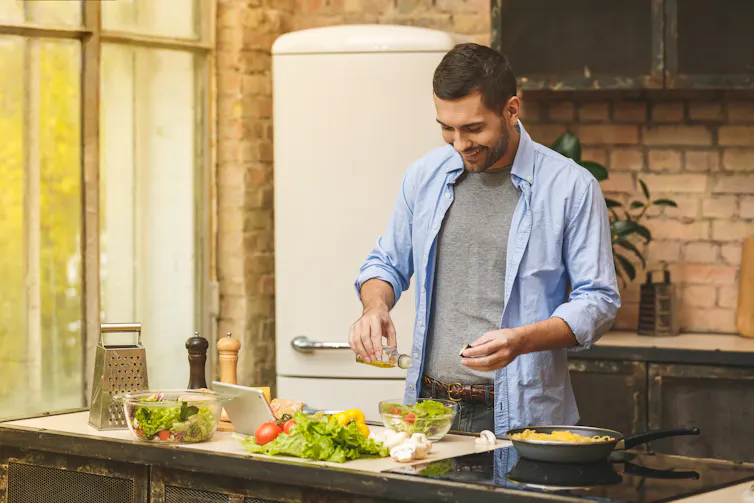 Young man in a blue shirt puts salt into a salad bowl while cooking in a kitchen.