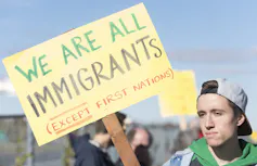 A man holds a sign that reads We Are All Immigrants (Except First Nations)