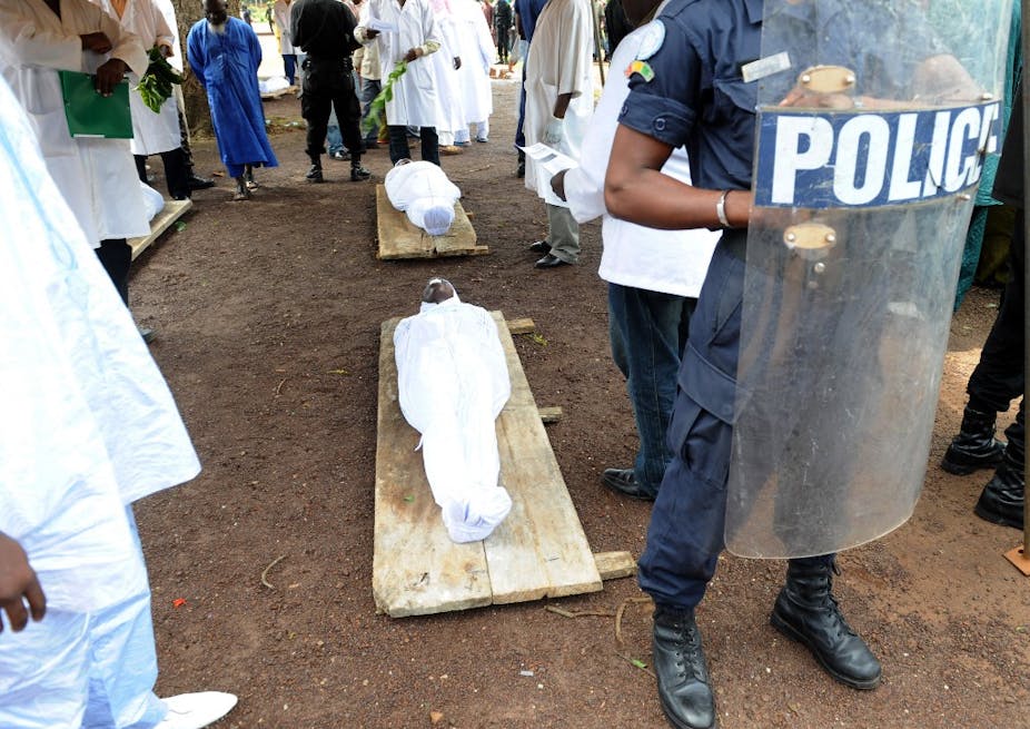 Corps dans des draps blancs sur des civières derrière un bouclier antiémeute siglé police.